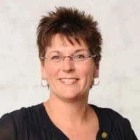 Portrait of a woman with short, spiky brown hair, wearing glasses, earrings, and a dark top, smiling in front of a neutral background.