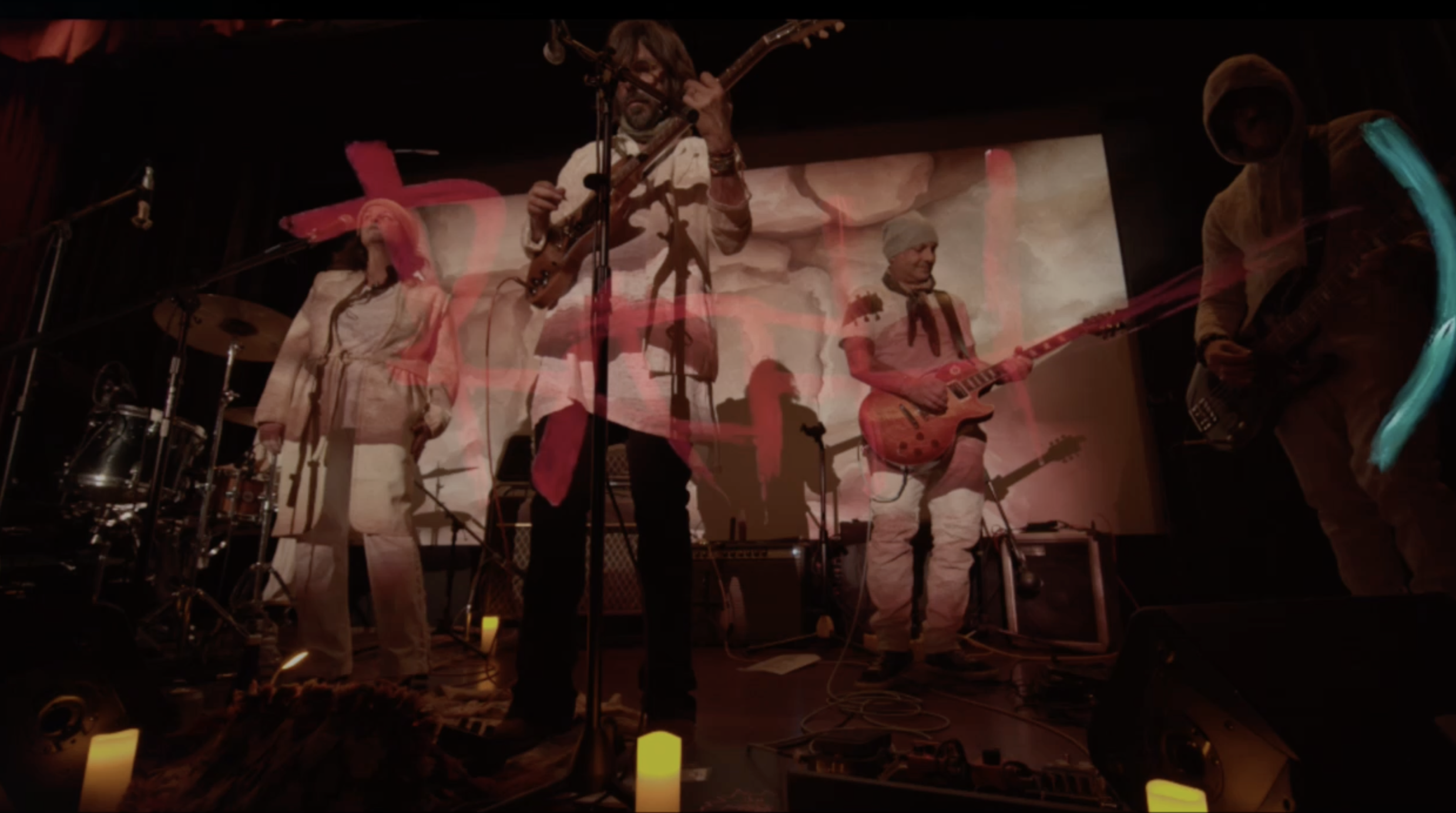 Rock band performing on stage with colorful abstract lighting and projections, featuring guitarists and a drummer.
