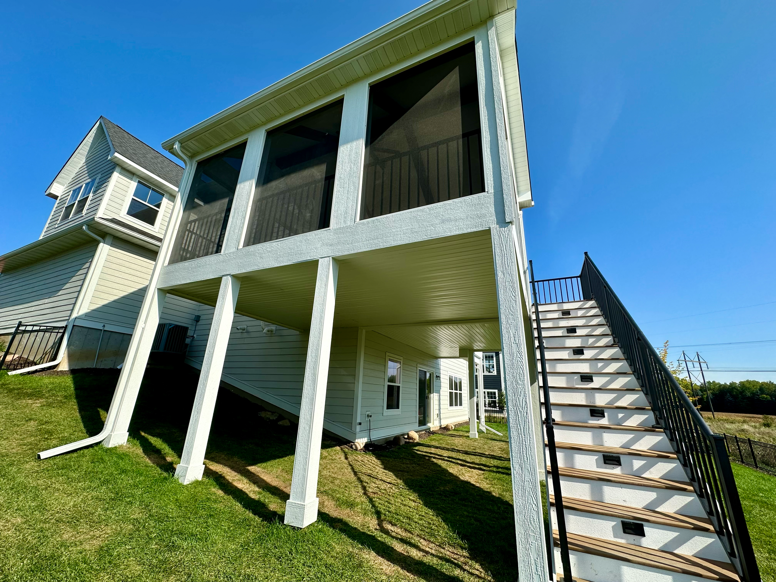Rear view of a modern house with a screened-in porch elevated on stilts, with stairs leading up to it, on a sunny day.