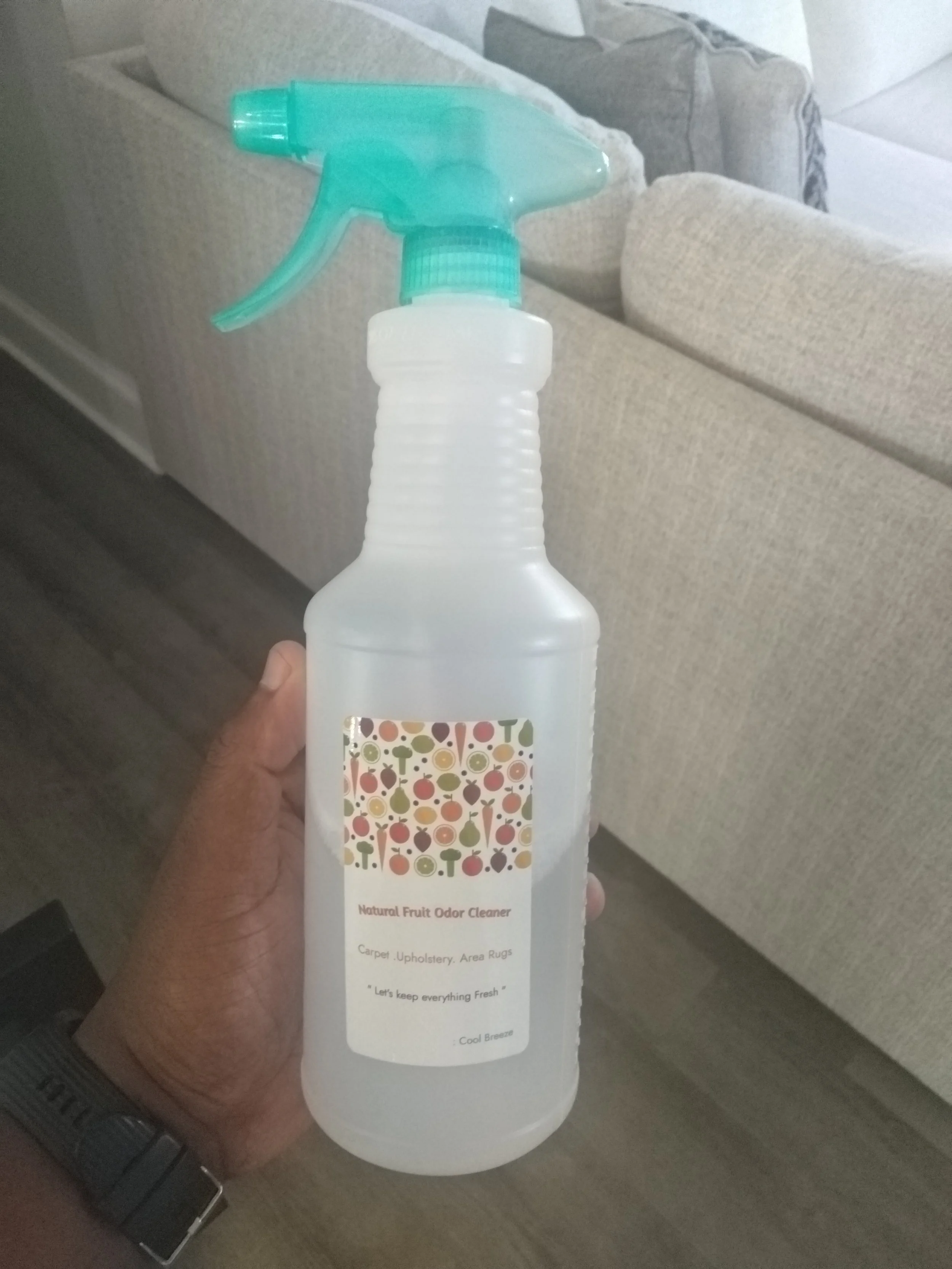 A person holding a spray bottle labeled 'Natural Fruit Odor Cleaner' with a colorful fruit pattern, designed for cleaning carpets, upholstery, and area rugs, against a background of a beige couch and a gray cushion.
