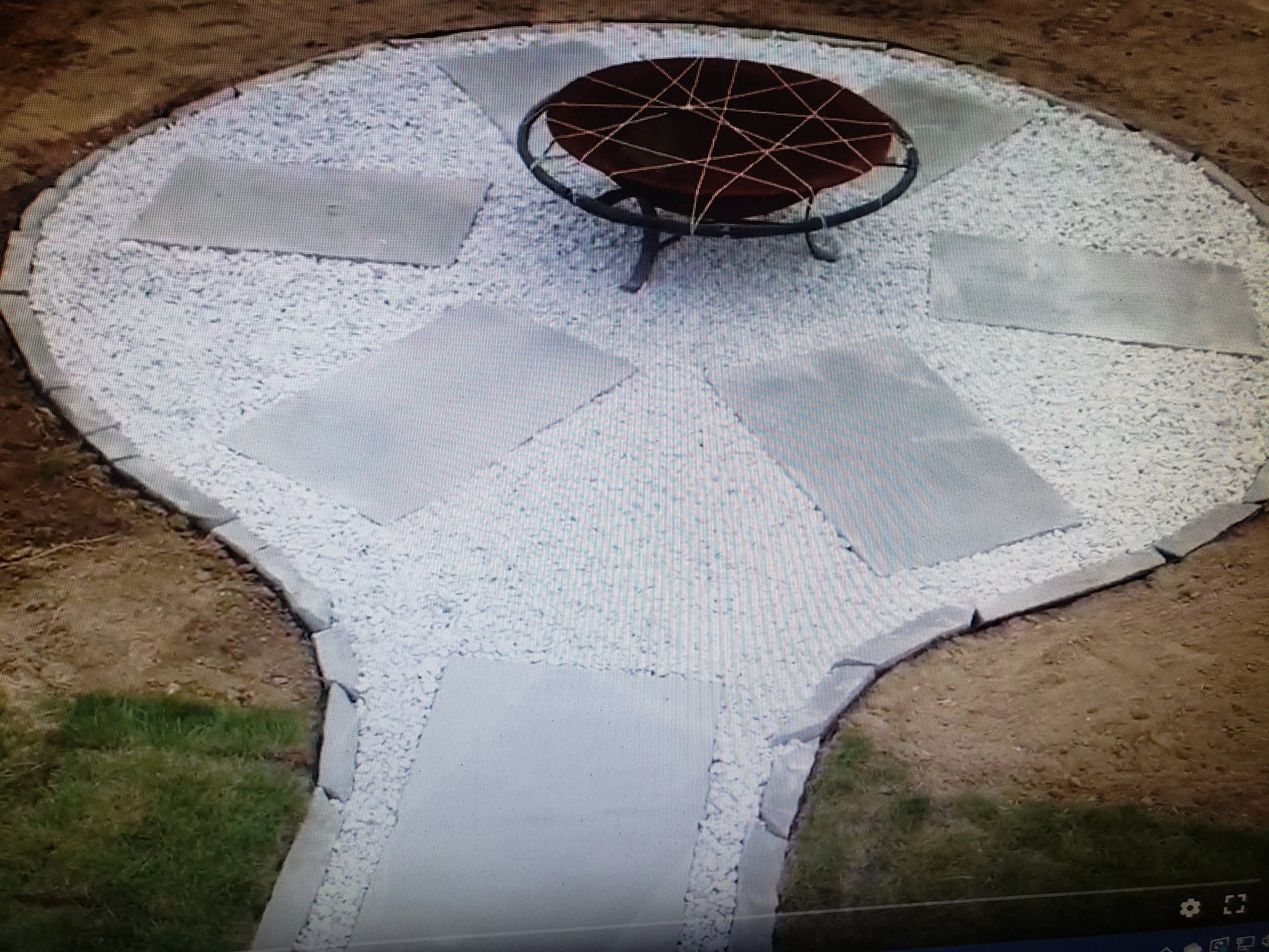 A yard with a circular patio area, a fire pit in the center, and a pathway made of concrete slabs surrounded by white gravel, with grass and dirt visible around the patio.