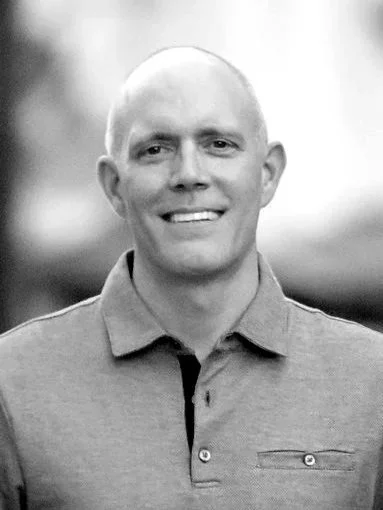 A smiling man with a clean-shaven face and bald head, wearing a polo shirt, outdoors in a black-and-white photo.