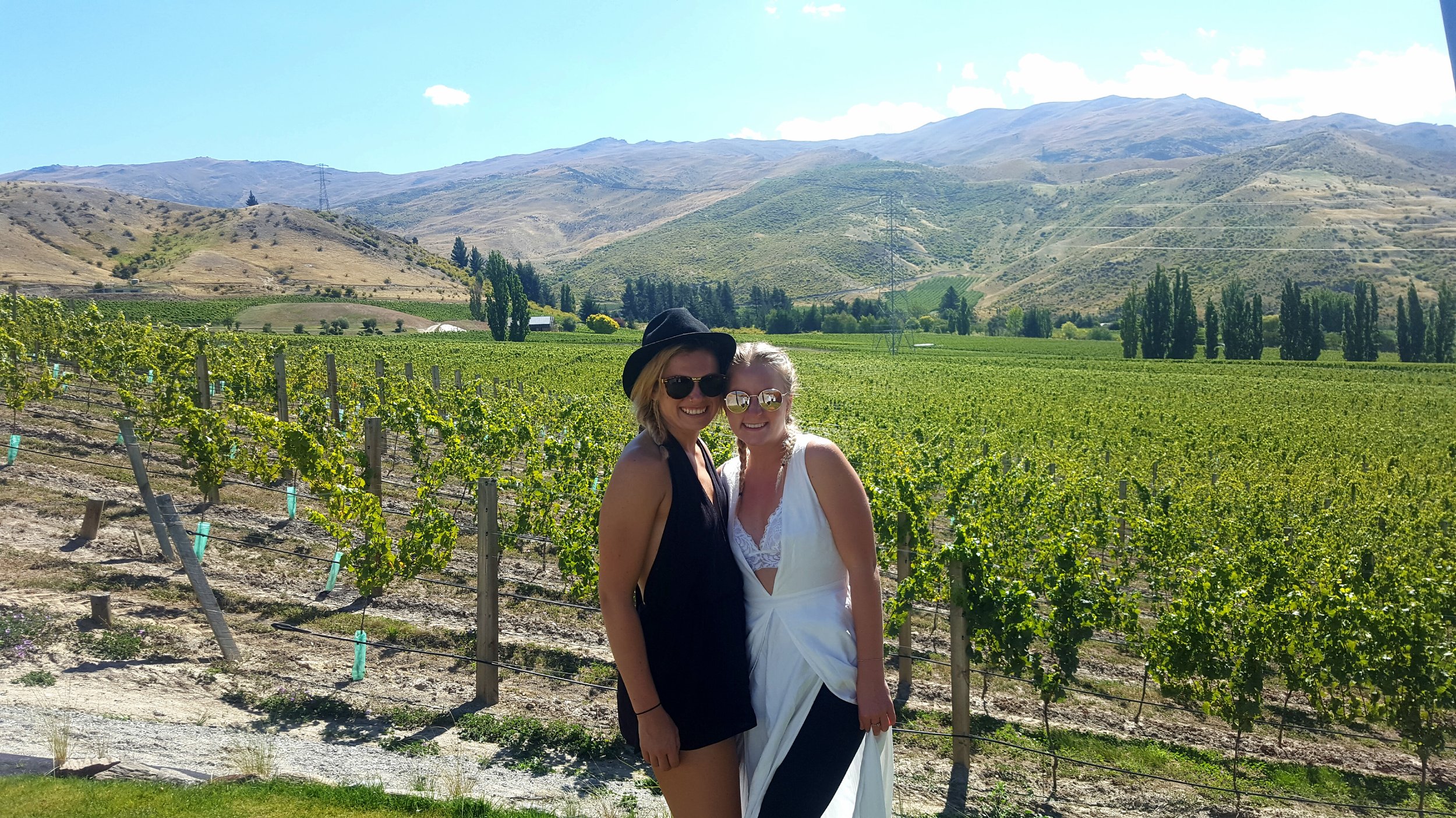 Two women in sunglasses standing in a vineyard with mountains in the background. https://www.centralotago.co.nz/