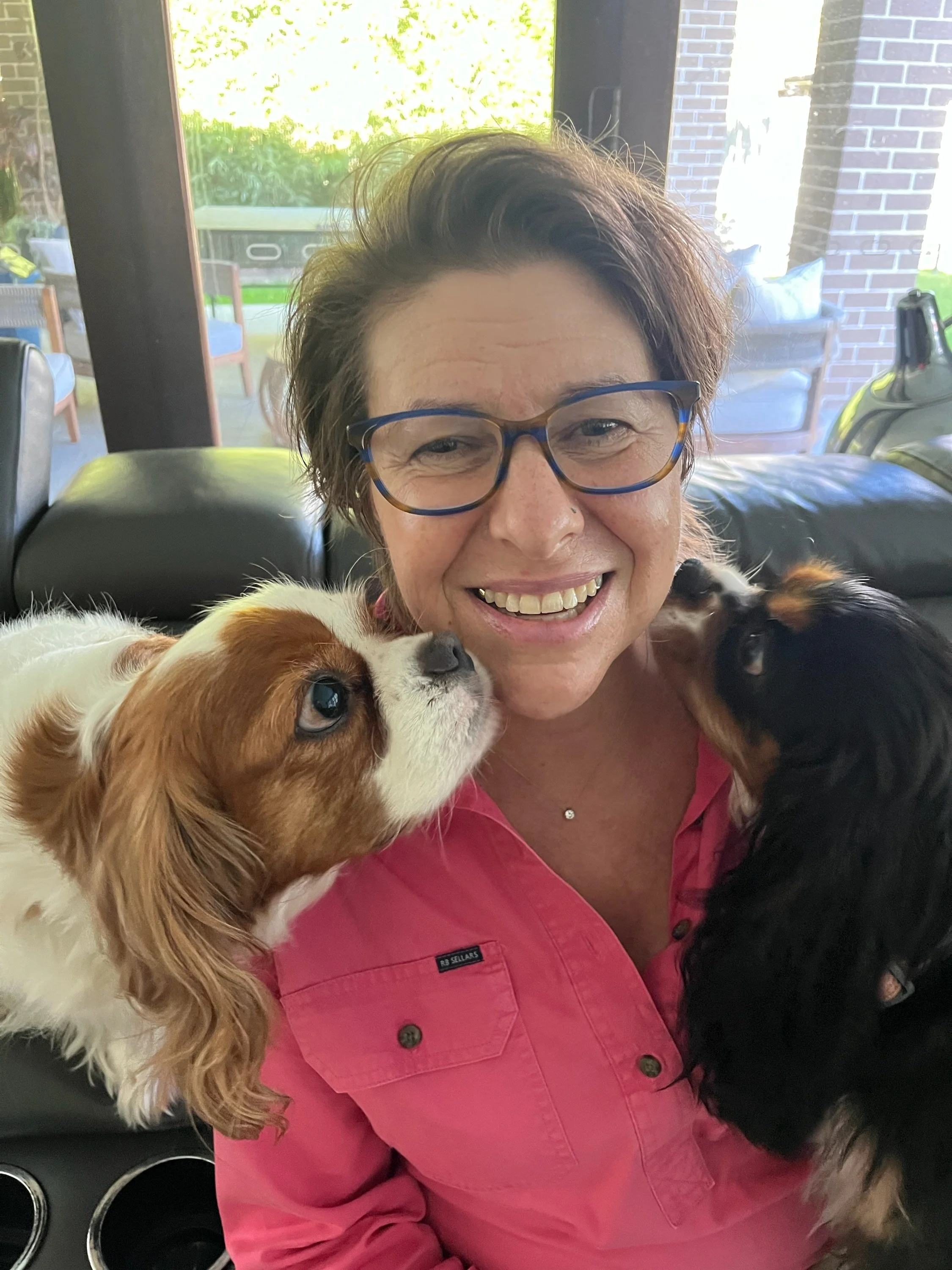 A woman with short brown hair, wearing glasses and a pink shirt, smiling as she holds two small dogs, one on each side of her face, inside a house.