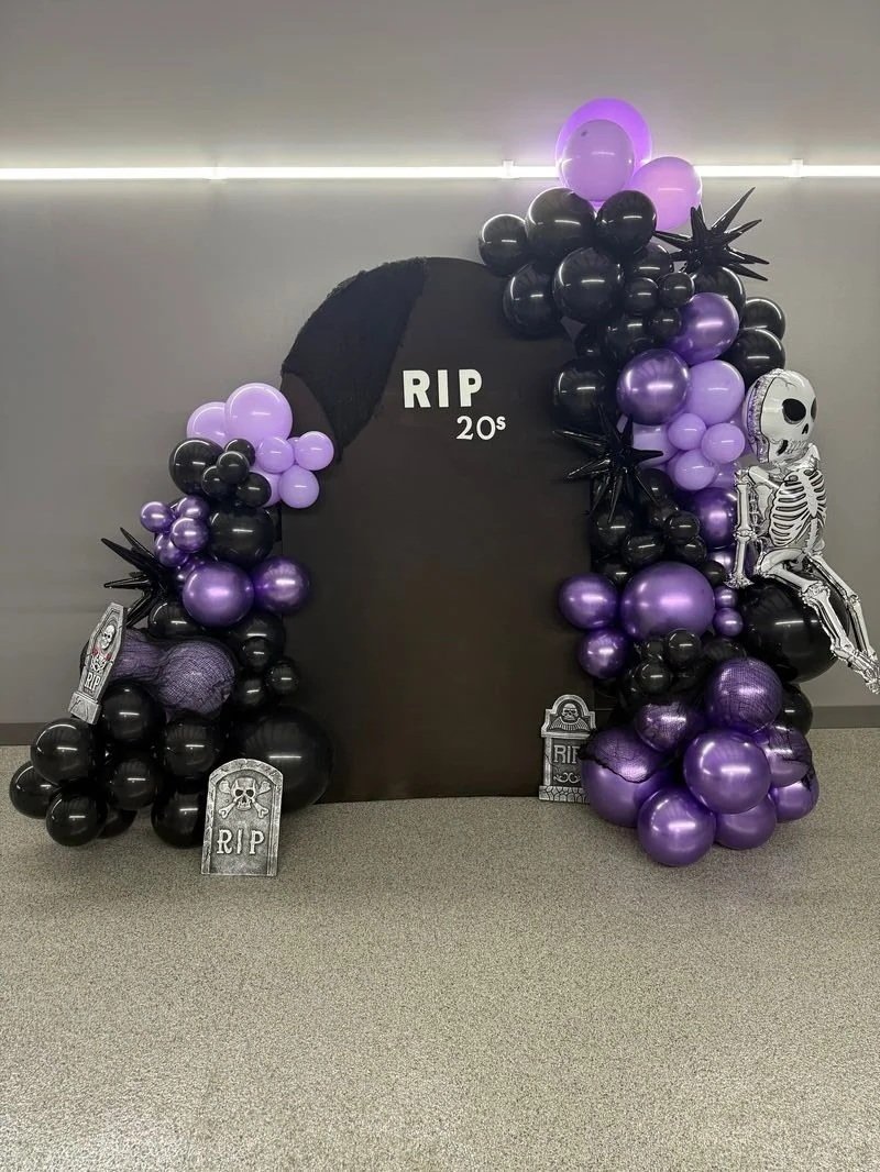 Halloween decoration with black and purple balloons, skeleton balloon, tombstone decorations, and a black arch with RIP and 20s text, set up for a spooky scene.
