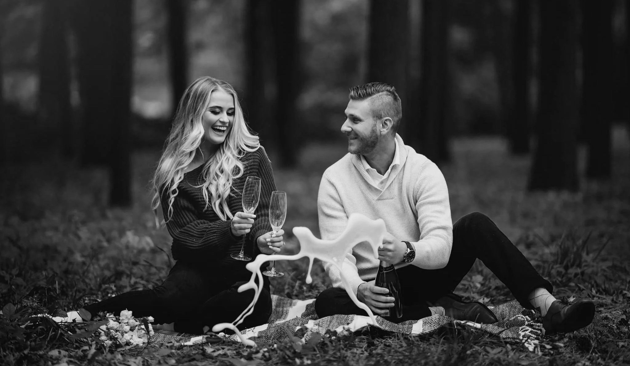 A black and white photo of a man and woman sitting on a blanket outdoors in a forest, smiling and holding glasses, with a decorative object and a bottle between them.