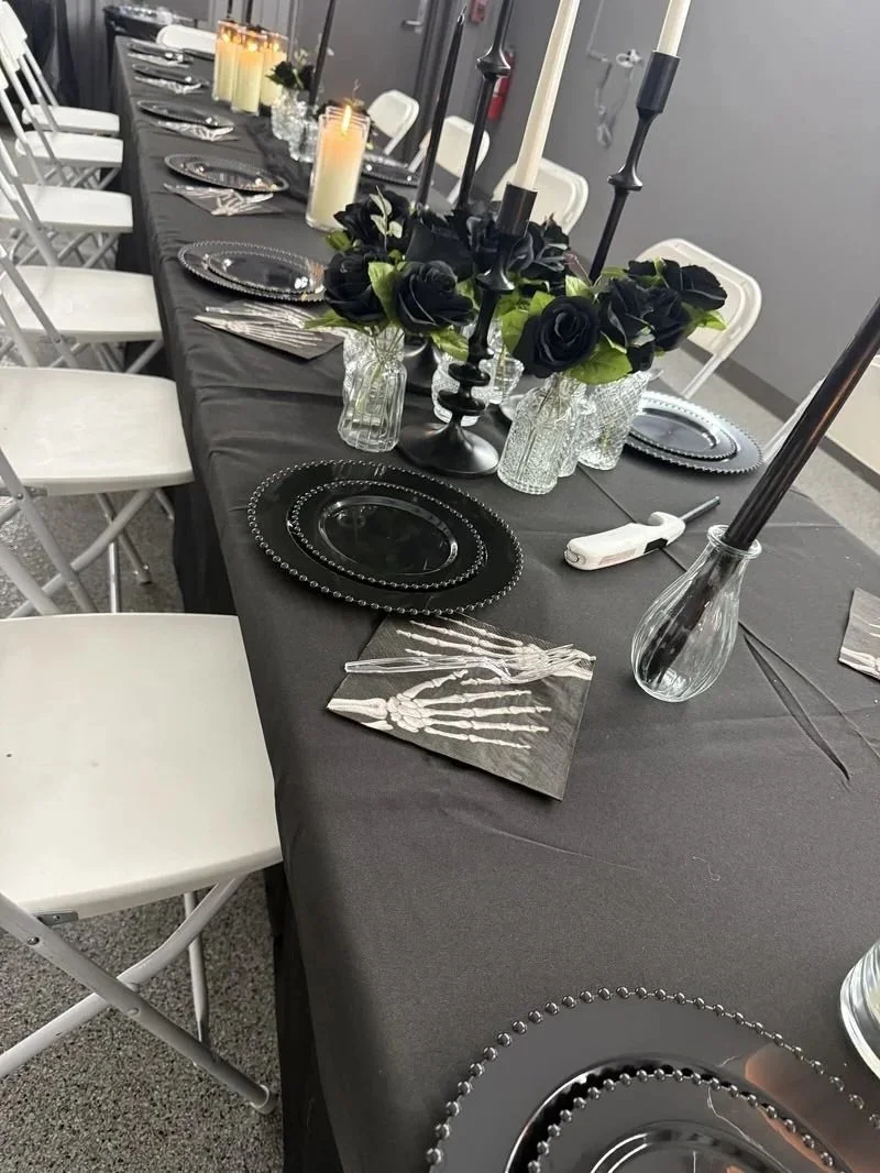 A long black table set for an event with silver and black decorations, black plates, black floral centerpieces, black candlesticks, and candles. The table has white chairs around it, with some candles lit in glass holders.