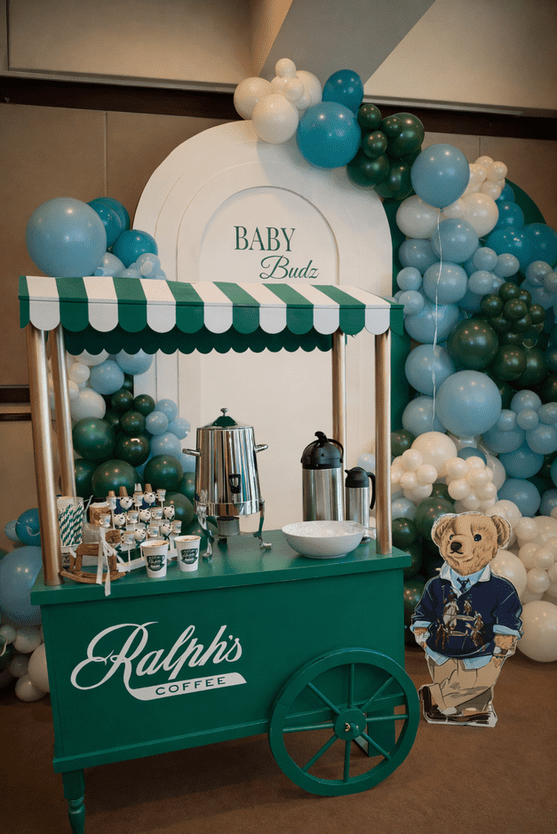 A coffee stand with a green and white striped canopy, labeled 'Ralph's Coffee,' in front of a balloon arch in shades of blue, white, and green. Behind the stand, a sign reads 'Baby Budz' and there is a cartoon cutout of a dog in clothing.