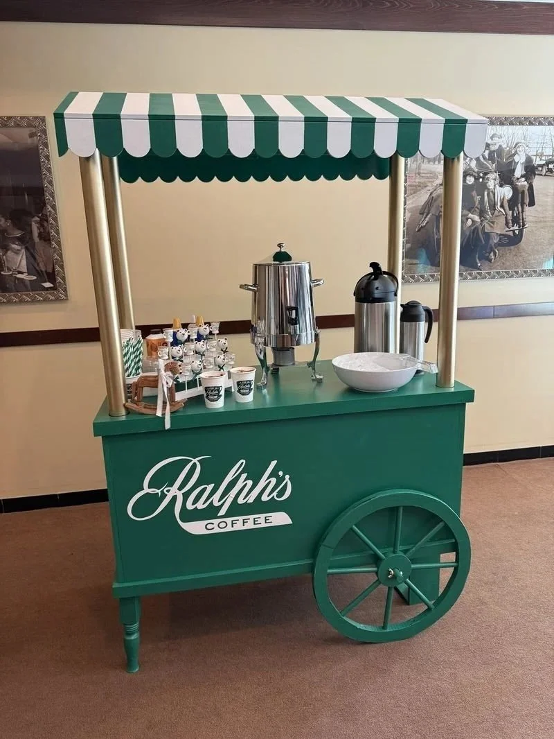 A green coffee cart with a striped green and white canopy and large wheel, branded "Ralph's Coffee". It has coffee brewing equipment, cups, and a large white bowl on top.