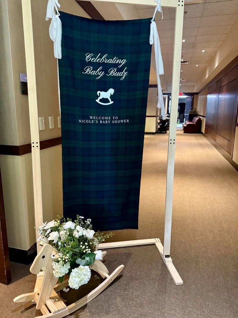 Sign for a baby shower reading 'Celebrating Baby Budz, Welcome to Nicole's Baby Shower,' with a rocking horse silhouette, and a wooden rocking horse planter with white flowers nearby.