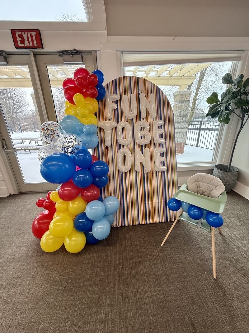 Colorful balloon arrangement with red, yellow, blue, and clear balloons next to a striped backdrop with white balloon letters spelling "FUN TO BE ONE"; a baby high chair with balloons attached; large window showing outdoor winter scene.