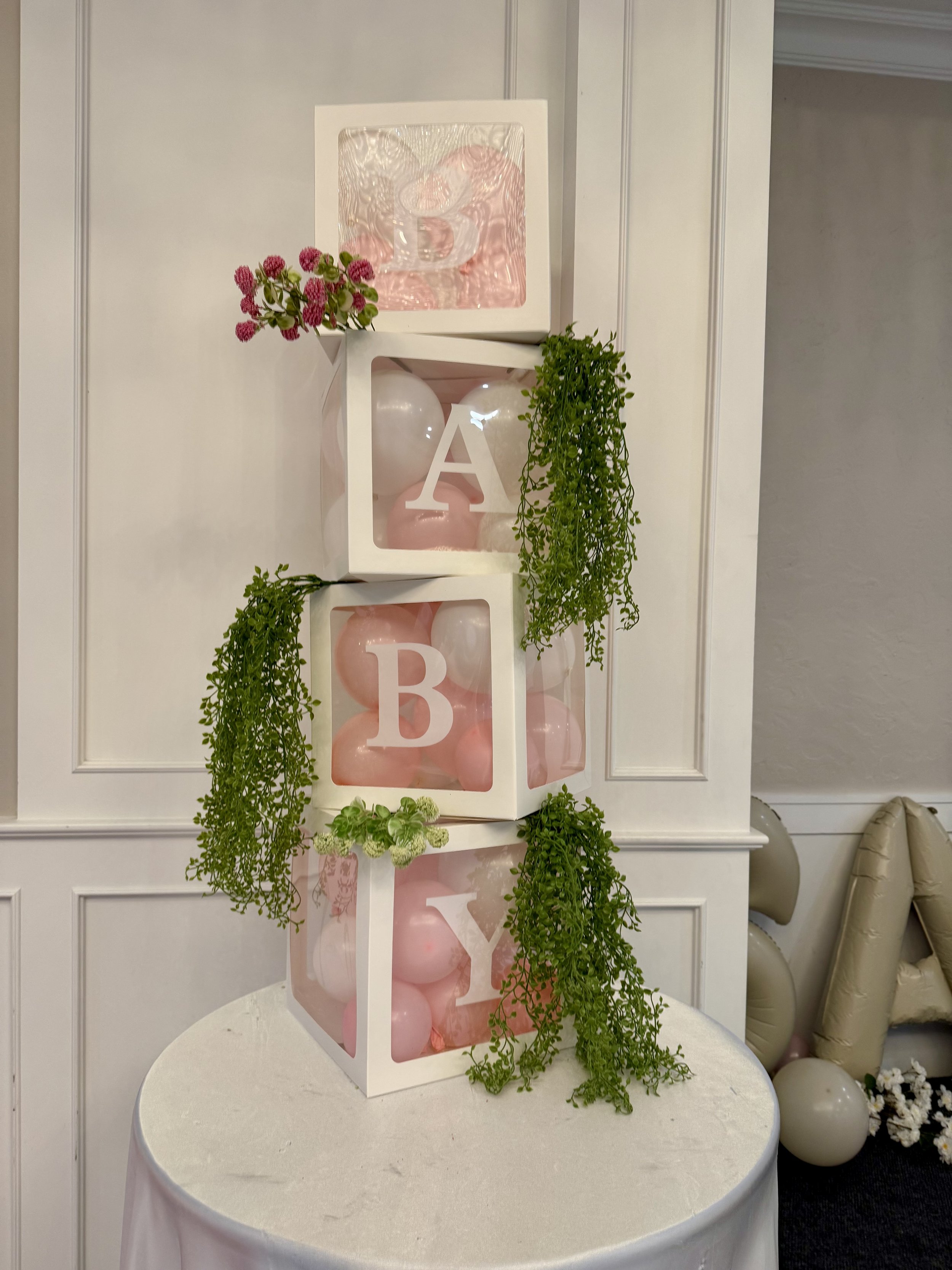 Decorative display with four white boxes containing pink and white balloons, each with a large white letter spelling 'BABY' and pink flowers and green leaves for decoration.