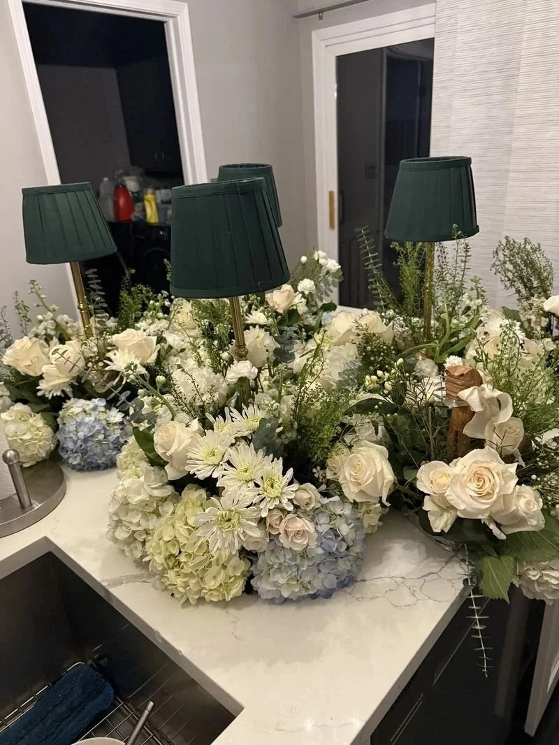 A kitchen countertop decorated with multiple arrangements of white and light blue flowers, including hydrangeas, roses, and daisies, with four small green lamps on top.