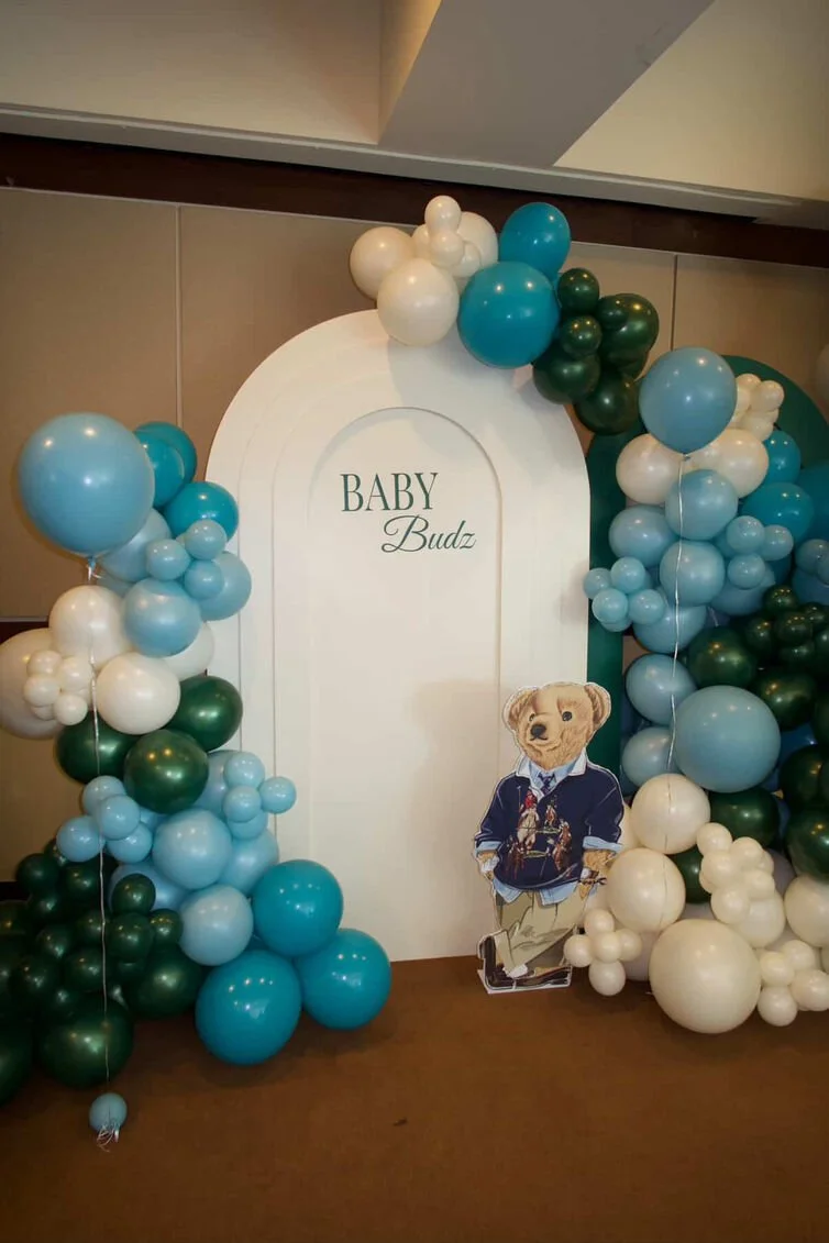 Decorative arch with blue, white, and green balloons around a sign that reads 'BABY Budz' with a cardboard cutout of a bear dressed in clothing.