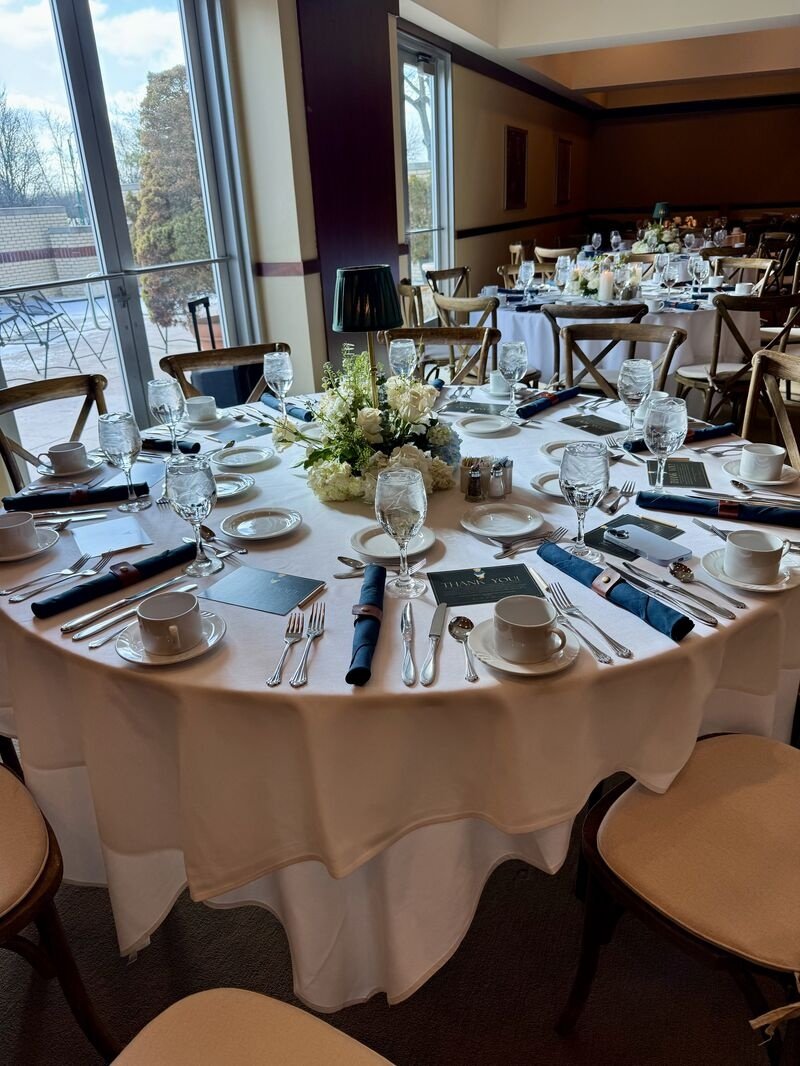 Round banquet table set for a formal event with white tablecloth, floral centerpiece, white dinnerware, glassware, and cloth napkins. Chairs are arranged around the table, and sunlight streams in through large windows.