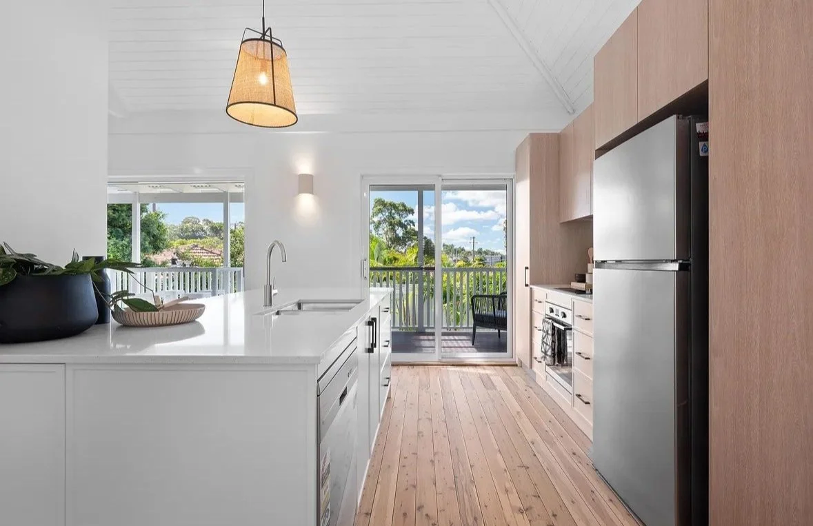 Modern kitchen with white countertops, stainless steel appliances, and light wood cabinetry. A pendant light hangs above the island, and a sliding door leads to a balcony with outdoor seating.