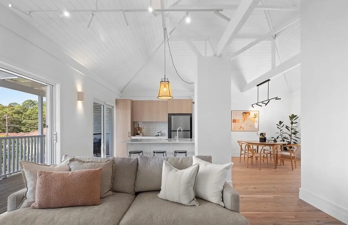 Modern open-plan living area with a beige sofa, high white ceilings, a kitchen with a stainless steel fridge, a dining area with wooden furniture, and a large window offering a view of trees outside.