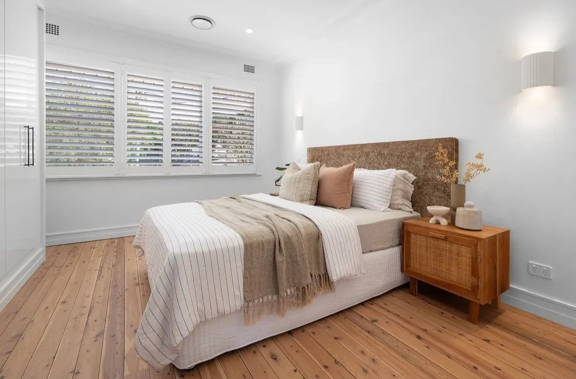 Modern bedroom with a bed, pillows, and wooden nightstand, featuring light wood flooring and white walls.