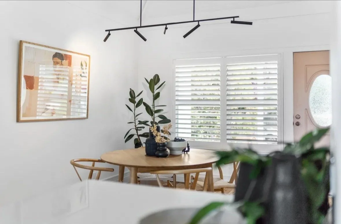 Modern dining room with round wooden table, chairs, large plant, wall art, track lighting, and window with blinds.