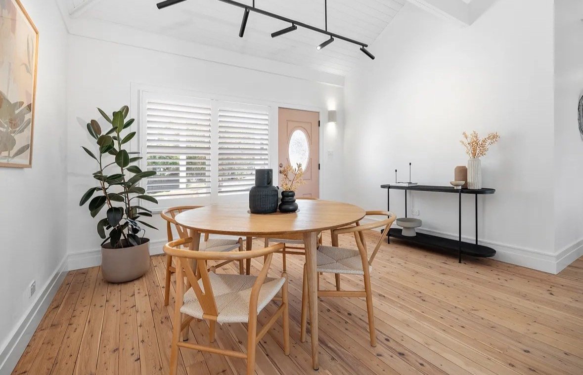 Minimalist dining room with a round wooden table, four chairs, potted plant, console table, and decorative items. Light wood flooring and white walls with window shutters and track lighting.