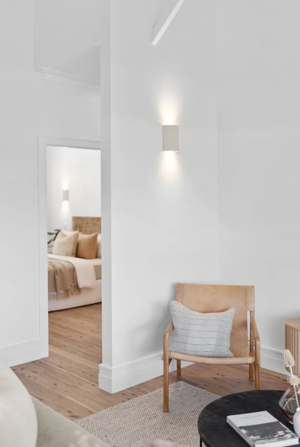 Minimalist living room with a wooden chair and pillow, white walls, and a doorway leading to a bedroom with a bed, pillows, and a wall light.