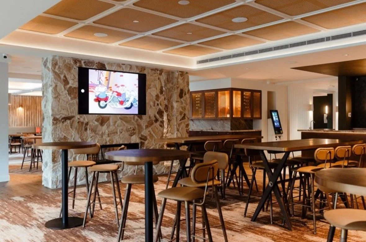 Modern lounge bar with high tables, chairs, stone wall, and TV displaying an animated show.