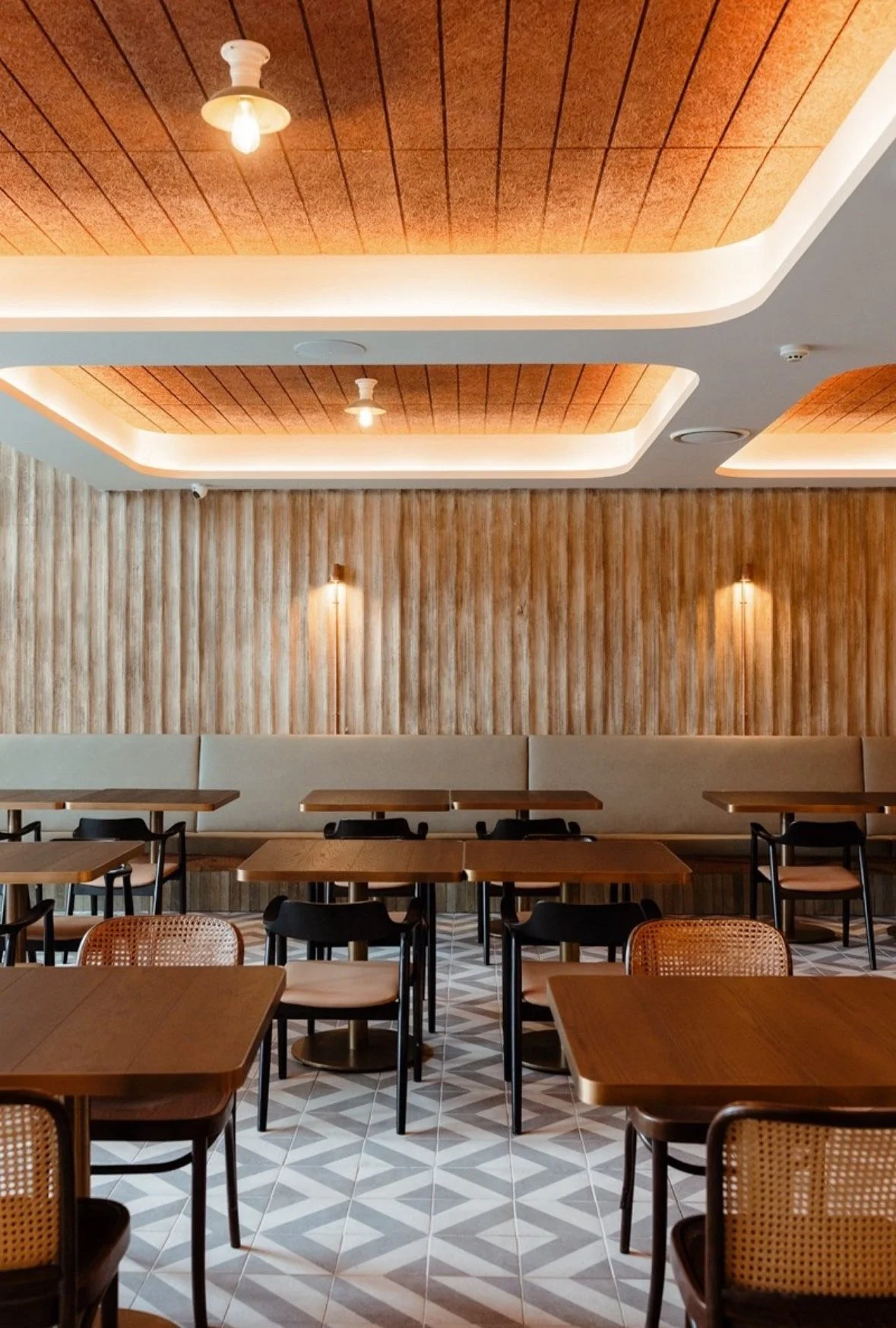 Modern restaurant interior with wooden tables and chairs, geometric floor tiles, wooden slatted walls, and ceiling lights.