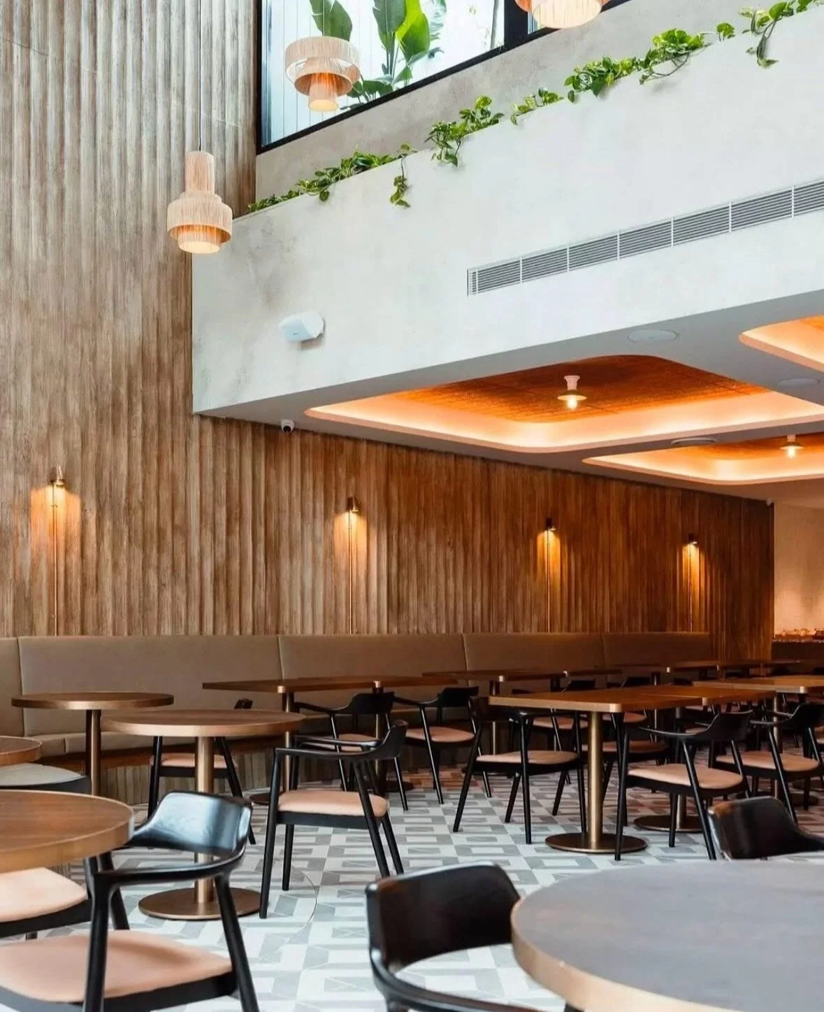 Modern restaurant interior with wooden accents, pendant lights, black chairs, and round tables. Vertical wooden wall panels and ceiling lights add warmth. Green plants are visible on a ledge above.