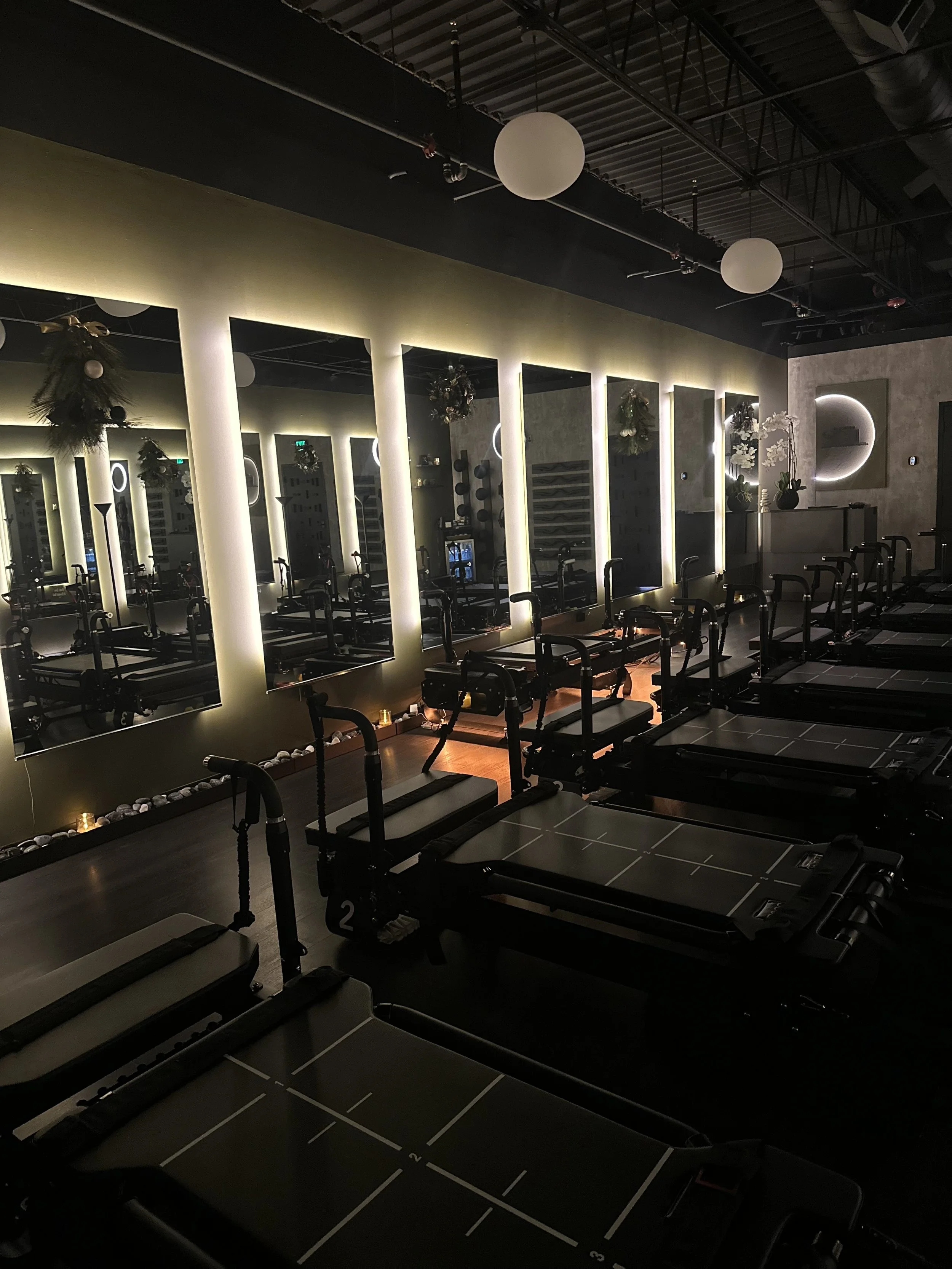 Interior of a Pilates or fitness studio with reformer Pilates machines, illuminated mirrors, and decorative wall lights, dimly lit for a relaxing atmosphere.