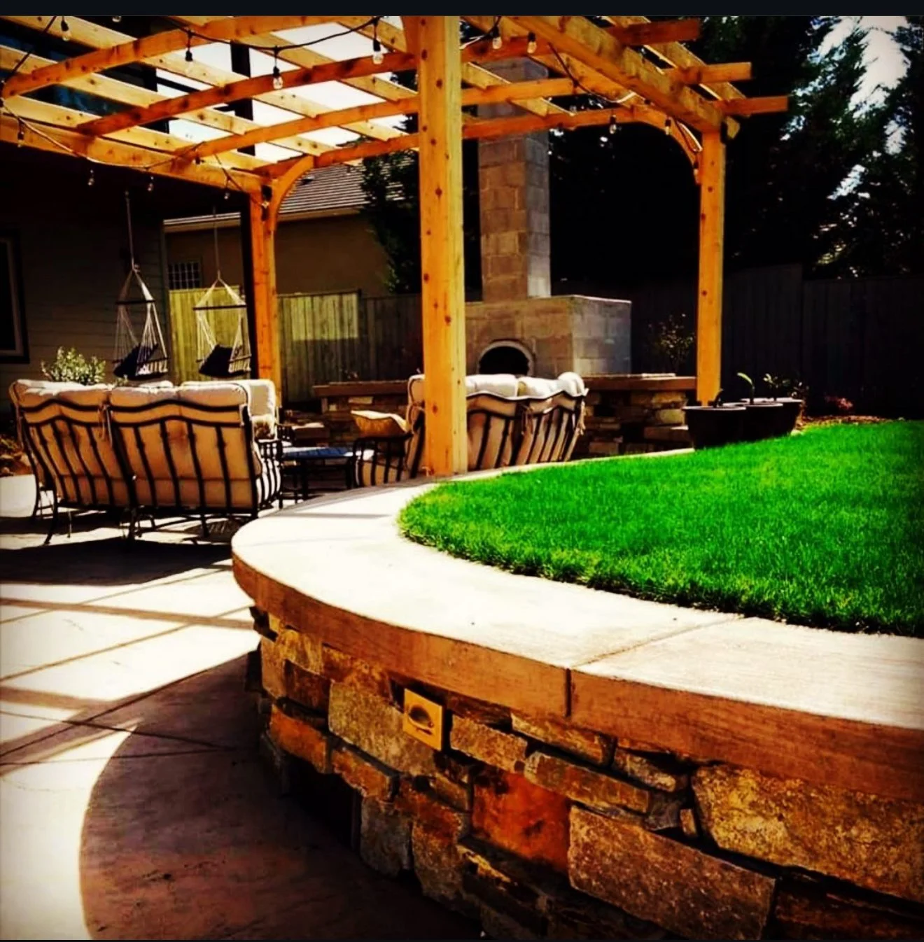 Backyard patio with wooden pergola, outdoor furniture, hanging swings, and a built-in brick fireplace, surrounded by a green grassy area and a wooden fence.