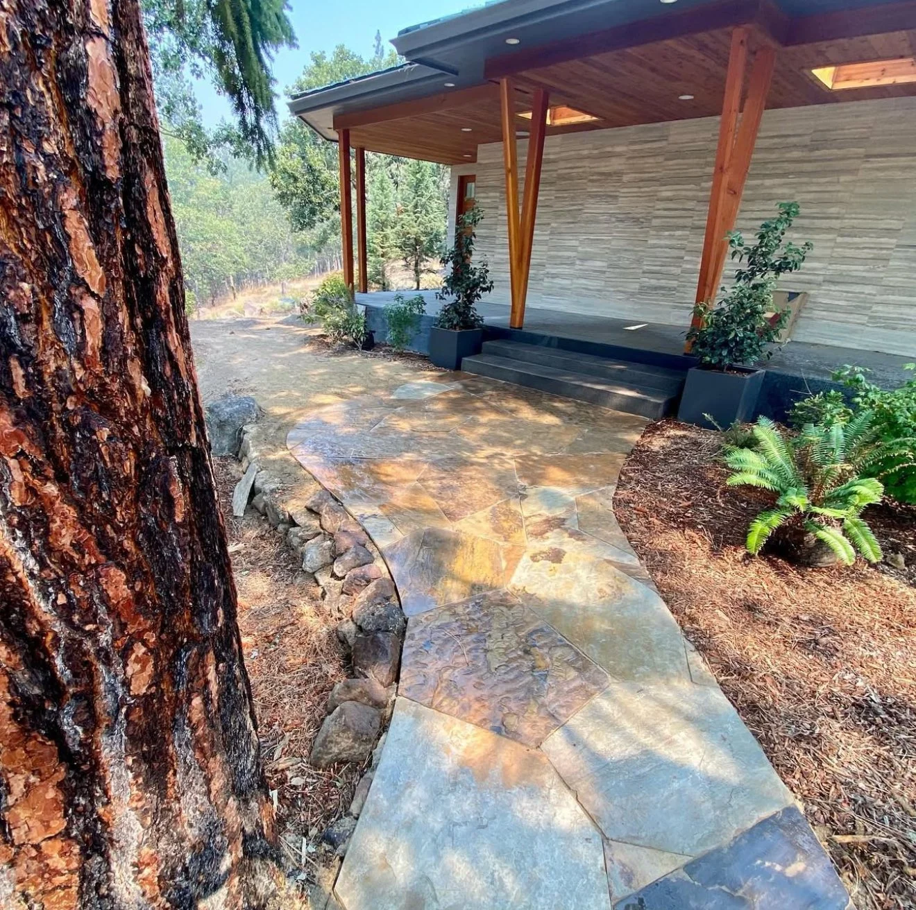 Stone pathway leading to a house with a covered porch, surrounded by trees and plants.