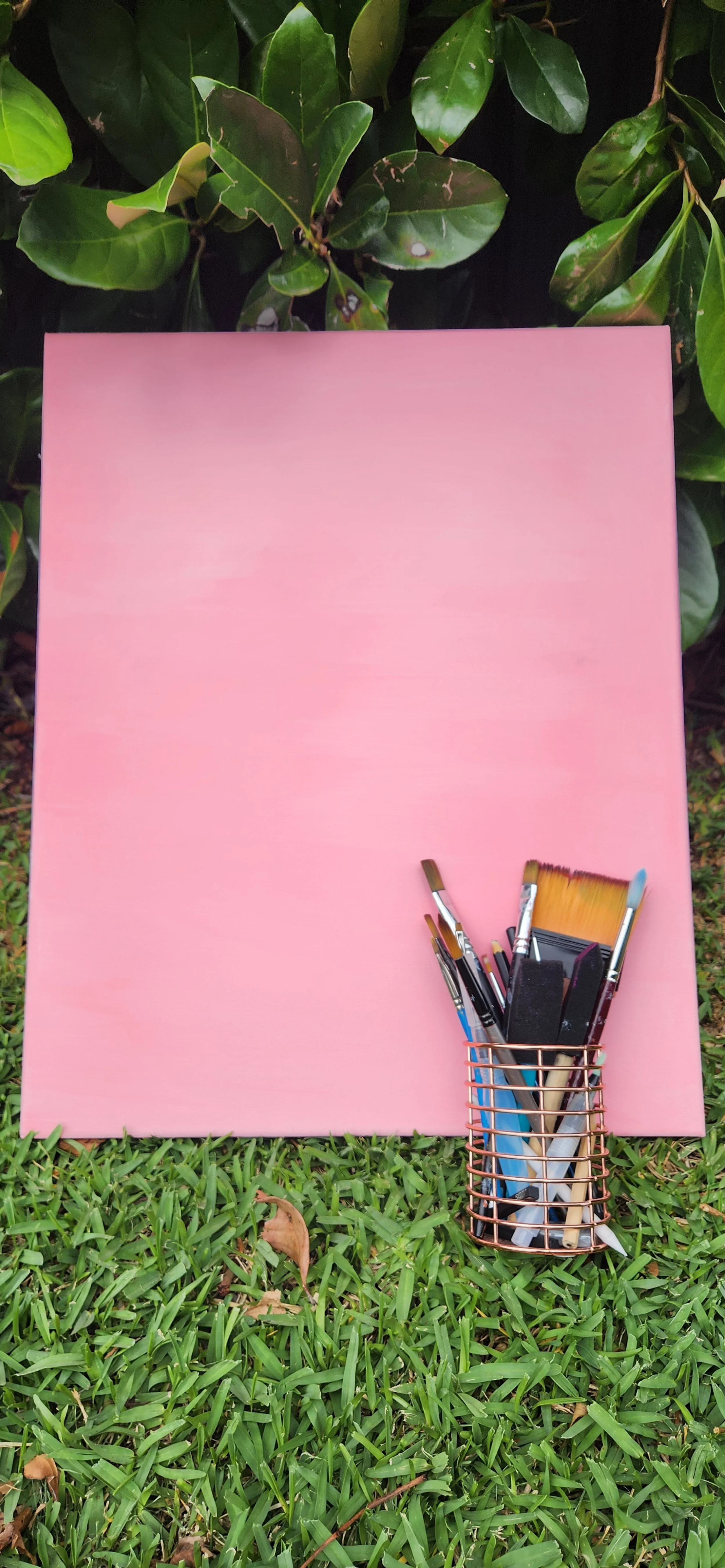 A pink canvas or board leaning against green bushes, with a copper wire basket holding various paintbrushes and painting tools in front of it, on green grass.