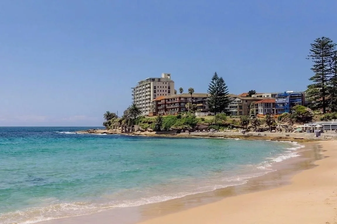 Beach with clear turquoise water, sandy shore, and buildings along the coastline under a blue sky.