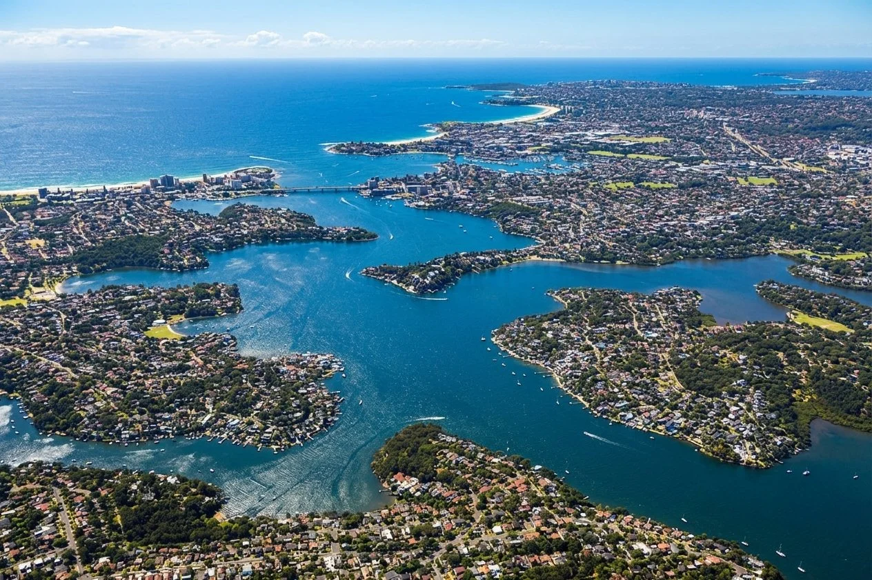 Aerial view of a coastal city with multiple waterways and islands, showing residential neighborhoods, green parks, and the ocean in the background.