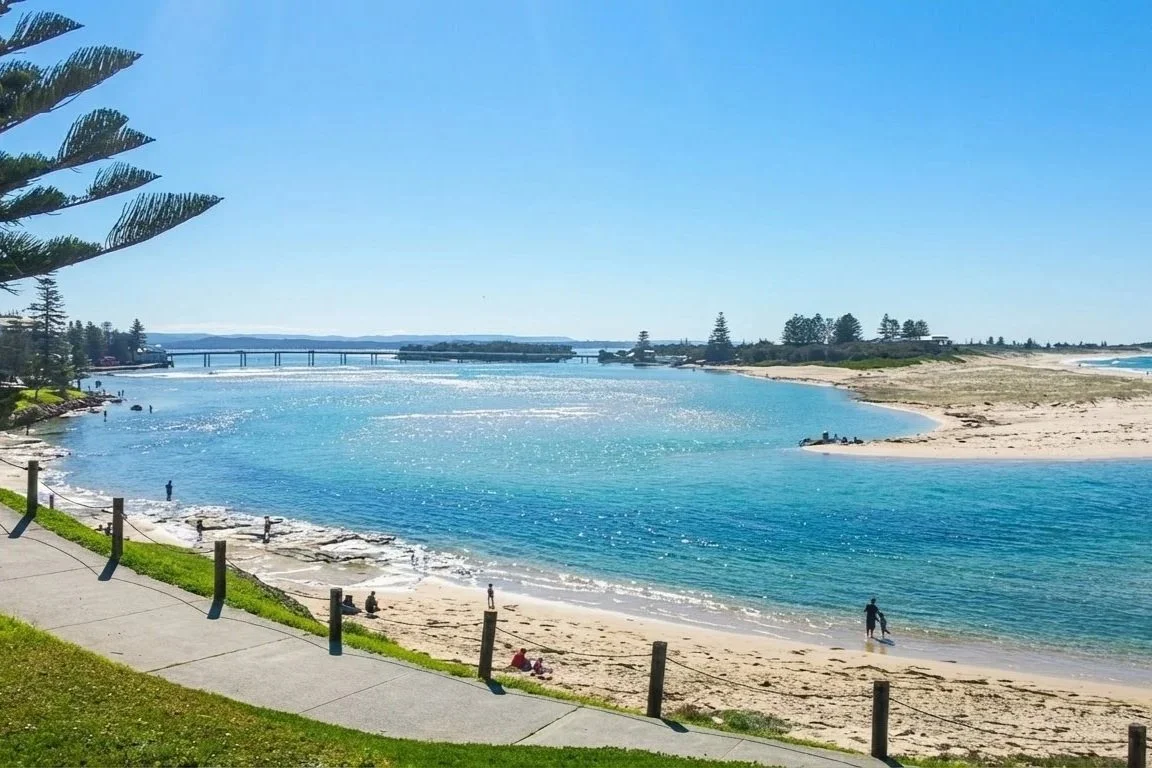A scenic view of a beach with clear blue water, sandy shores, and a few people walking or sitting on the beach, with a grassy area and trees in the foreground and a bridge in the background under a bright blue sky.