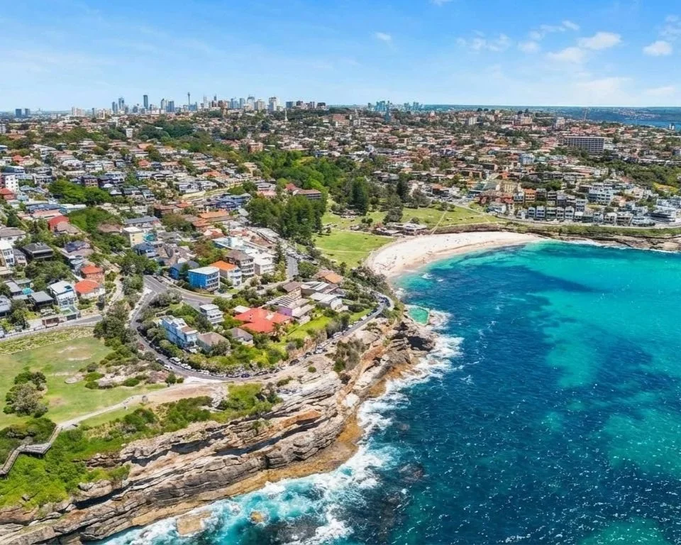 Aerial view of a coastal city with residential areas, a sandy beach, and an ocean with waves crashing against rocky cliffs under a partly cloudy sky.