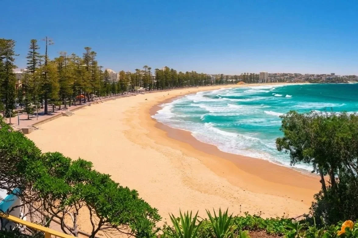 A sandy beach with turquoise waves crashing onto the shore, lined with trees and a promenade with benches and streetlights, under a clear blue sky.