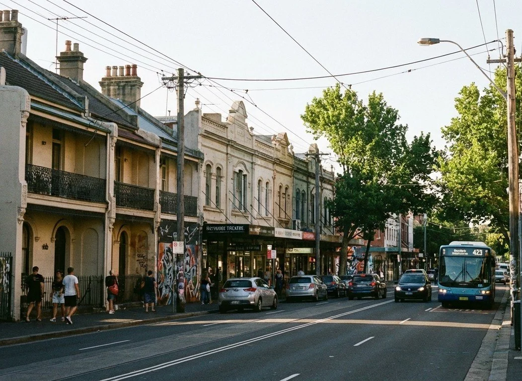 Street view with cars parked along the curb and pedestrians walking on the sidewalk, featuring historic-style buildings, utility poles with overhead wires, and a blue bus on the right side of the road under trees.
