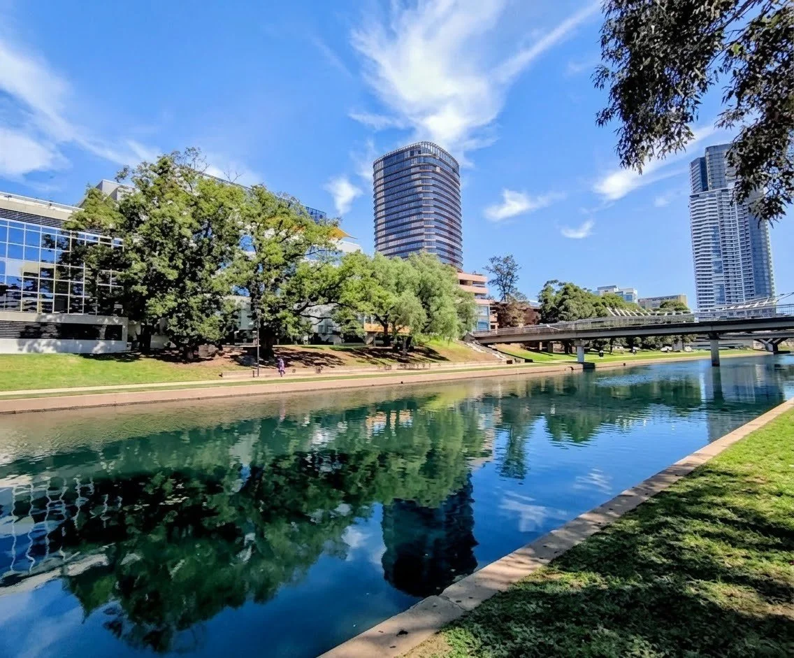 Cityscape with high-rise buildings, trees, a river, and a blue sky with clouds.