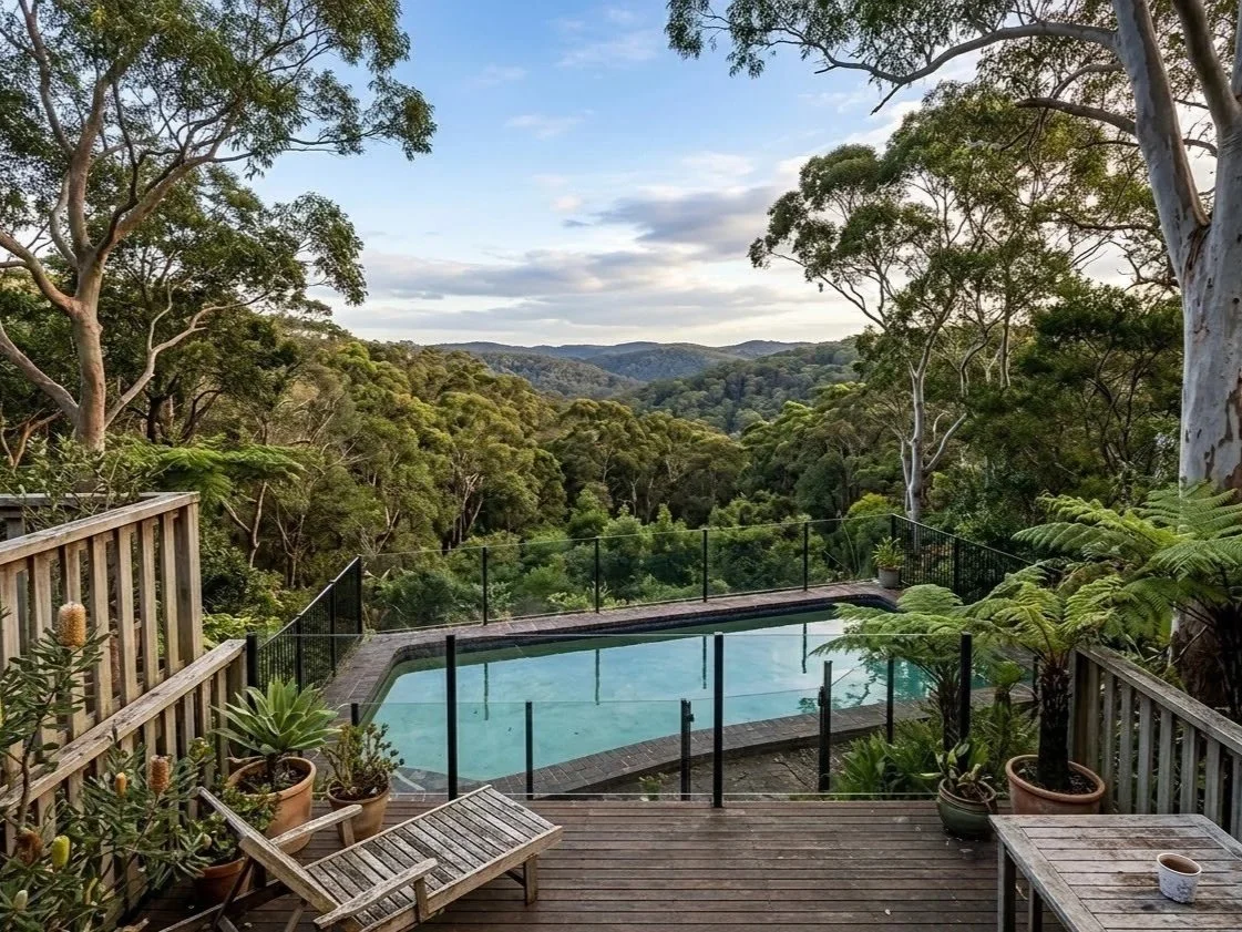 A backyard swimming pool on a wooden deck surrounded by potted plants, overlooking a forested landscape with rolling hills and trees under a partly cloudy sky.