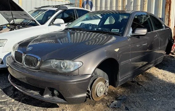 A gray BMW car with missing front wheels, parked outdoors on rocky ground next to other vehicles.