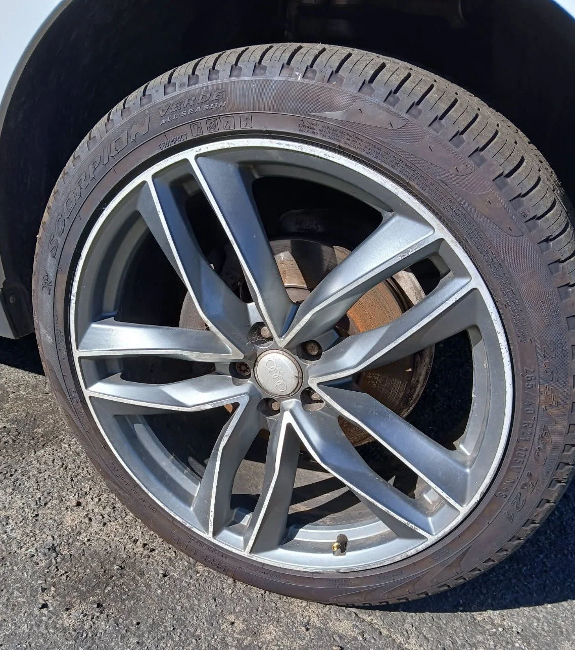 Close-up of a AUDI car wheel with a silver rim and an all-season tire, mounted on a vehicle parked on a gravel surface.