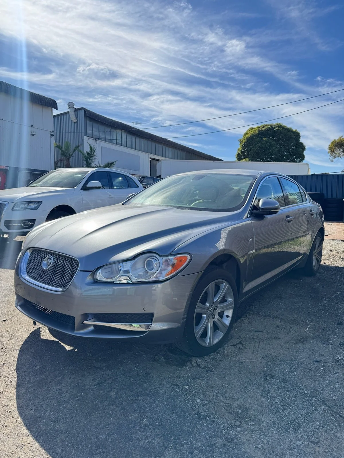 A silver luxury sedan car parked on a gravel lot during daytime, with blue skies and a few clouds overhead. Other vehicles and buildings are visible in the background.