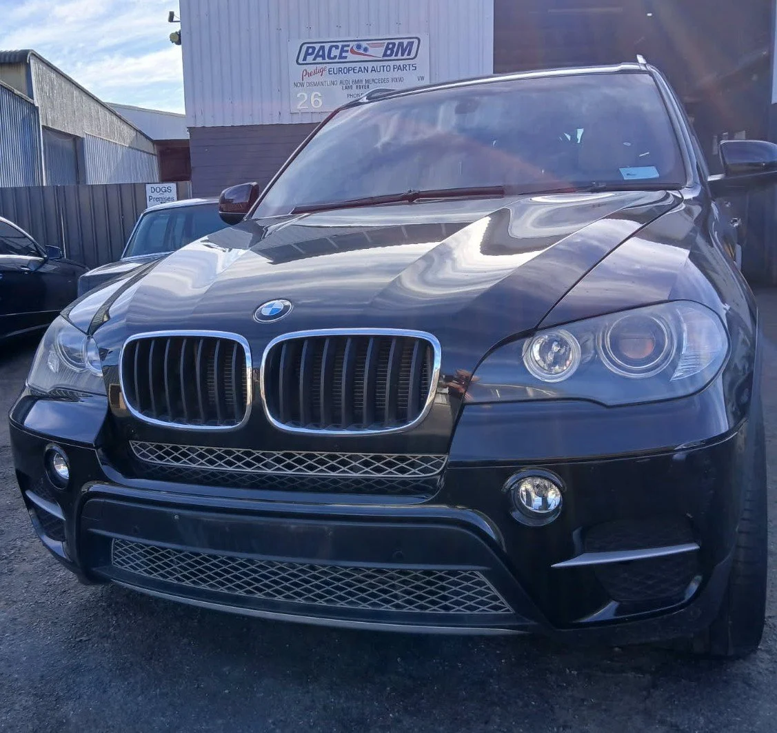Black BMW SUV parked at a dealership lot with a sign for European auto parts in the background.