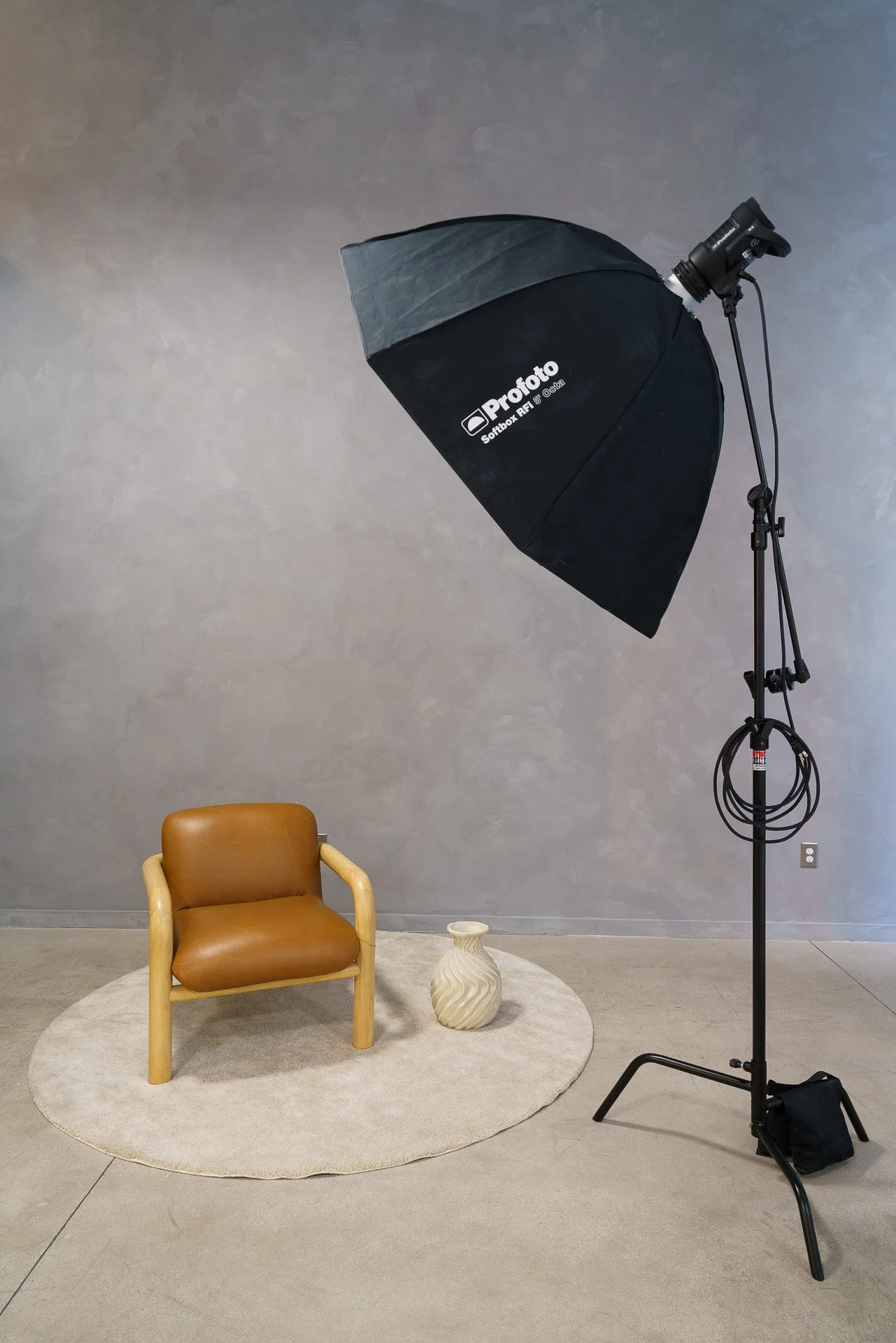 A professional photoshoot setup with a large black softbox light, a brown armchair on a light gray circular rug, a small decorative vase, and a gray wall background, indicating a studio interview or portrait session.