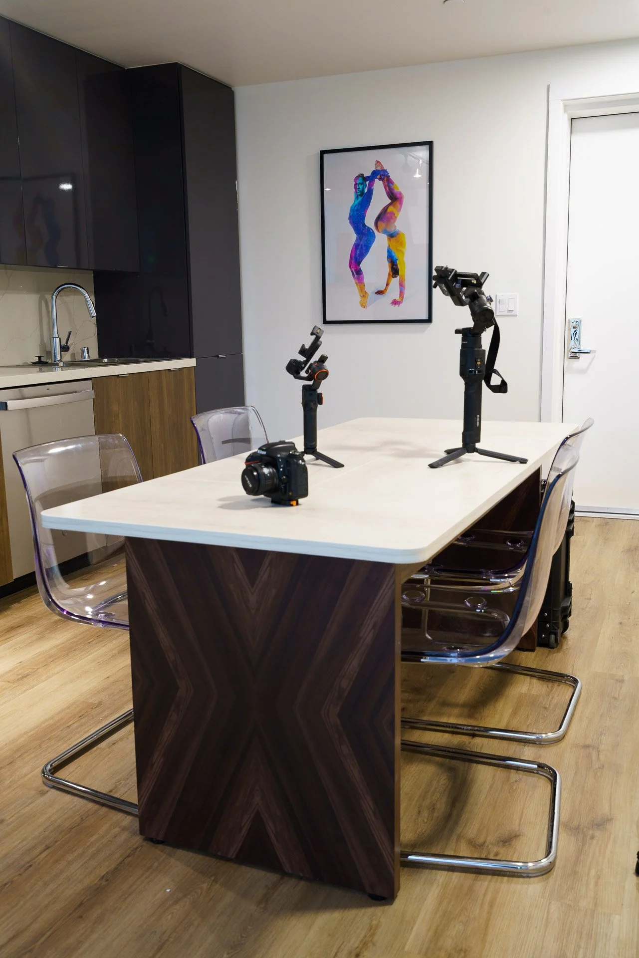 A modern kitchen with black cabinets, a sink, a white countertop, and a wooden cabinet. In the foreground, a white table with five transparent chairs and various camera equipment, including three camera stabilizers and a camera, placed on the table. On the wall, there is a colorful art print of two women performing acrobatic poses.