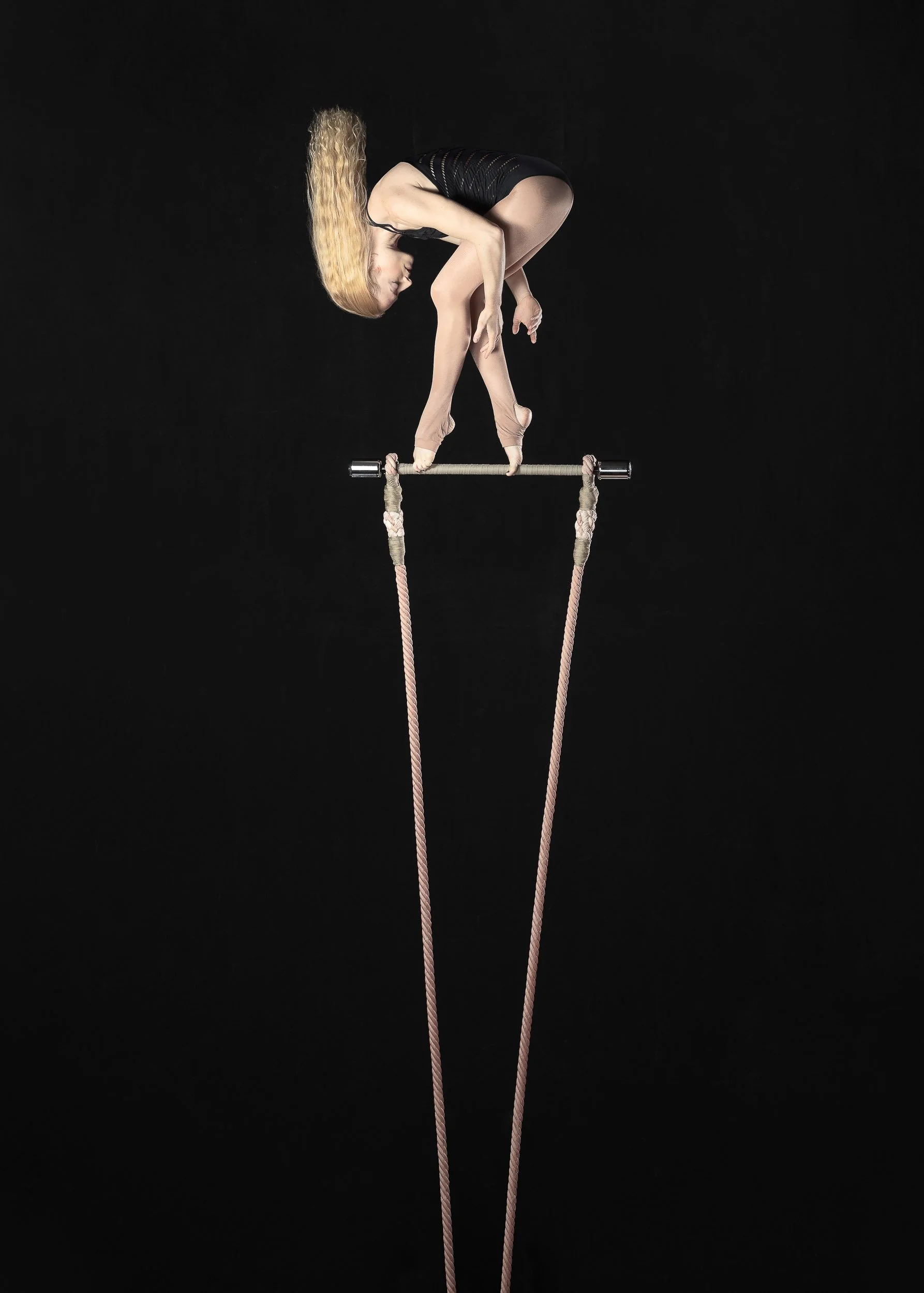 A female gymnast is balancing on a narrow beam, with her arms extended downward and her back arched, set against a black background.