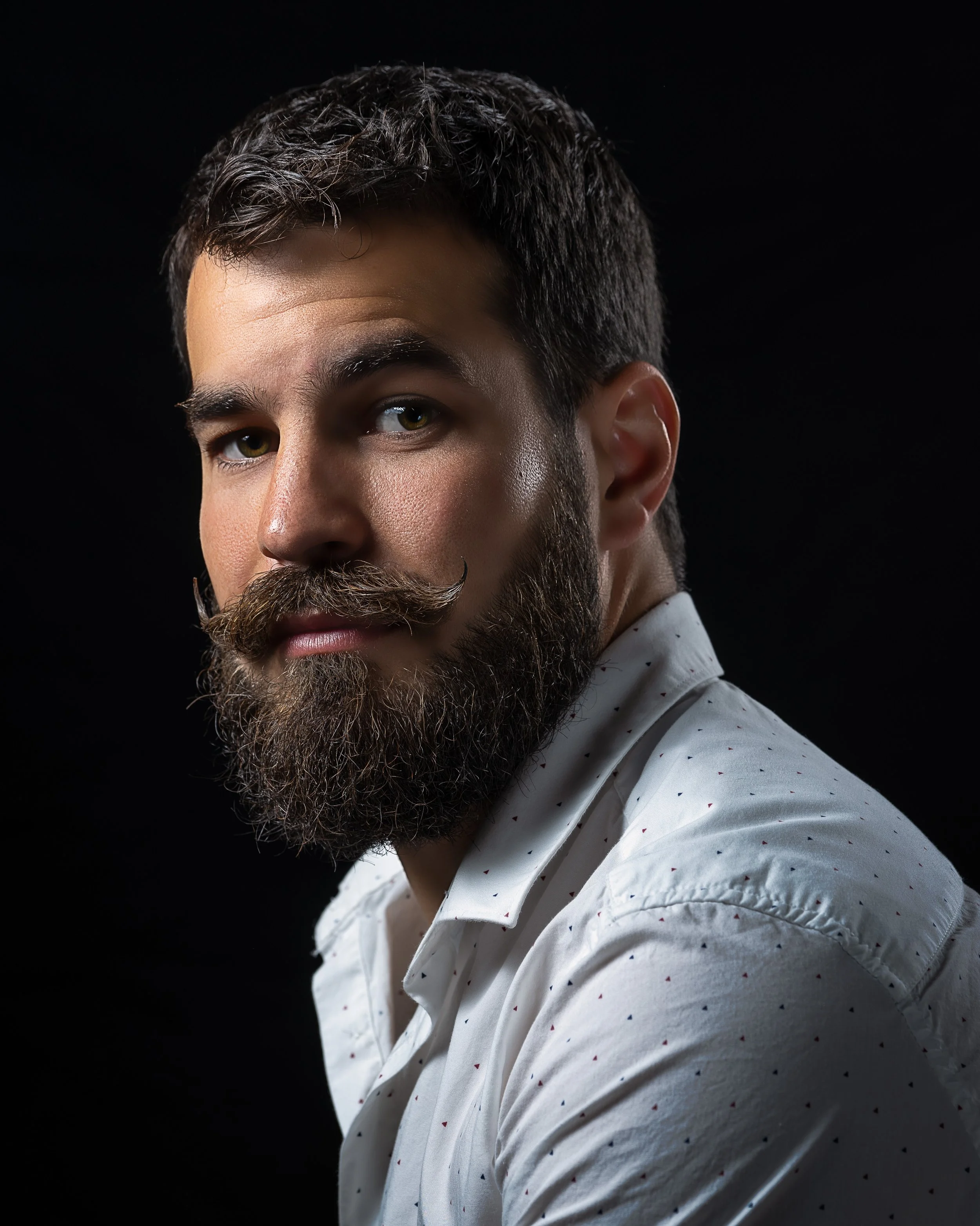 Close-up of a man with a beard and mustache, wearing a white shirt with small colorful patterns, against a black background.