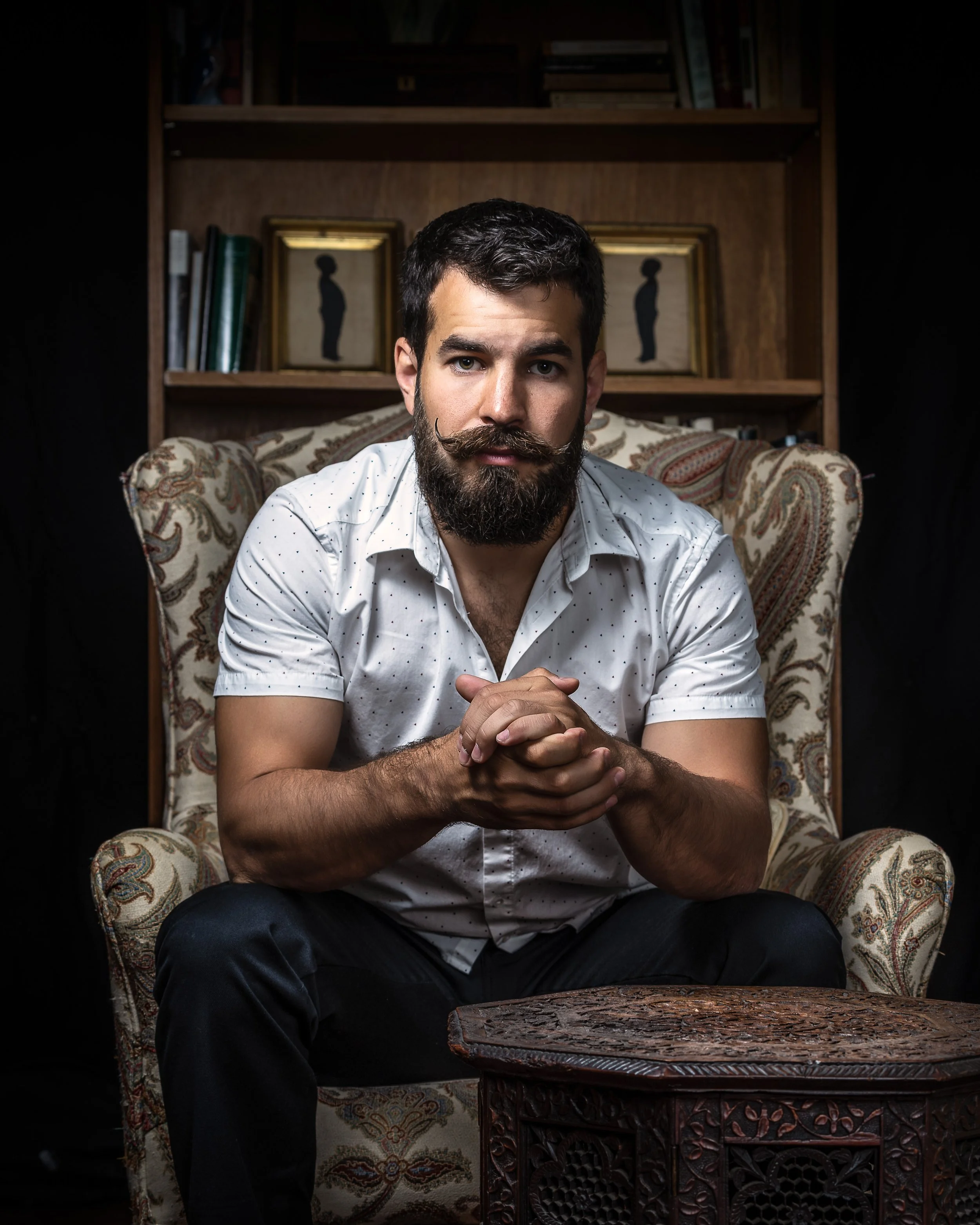A young man with a beard and mustache, wearing a white shirt with small polka dots, sitting on an ornate armchair with a floral pattern. He has his hands clasped together on his lap, looking directly at the camera with a serious expression. Behind him is a wooden bookshelf with books and framed silhouettes of a standing woman.