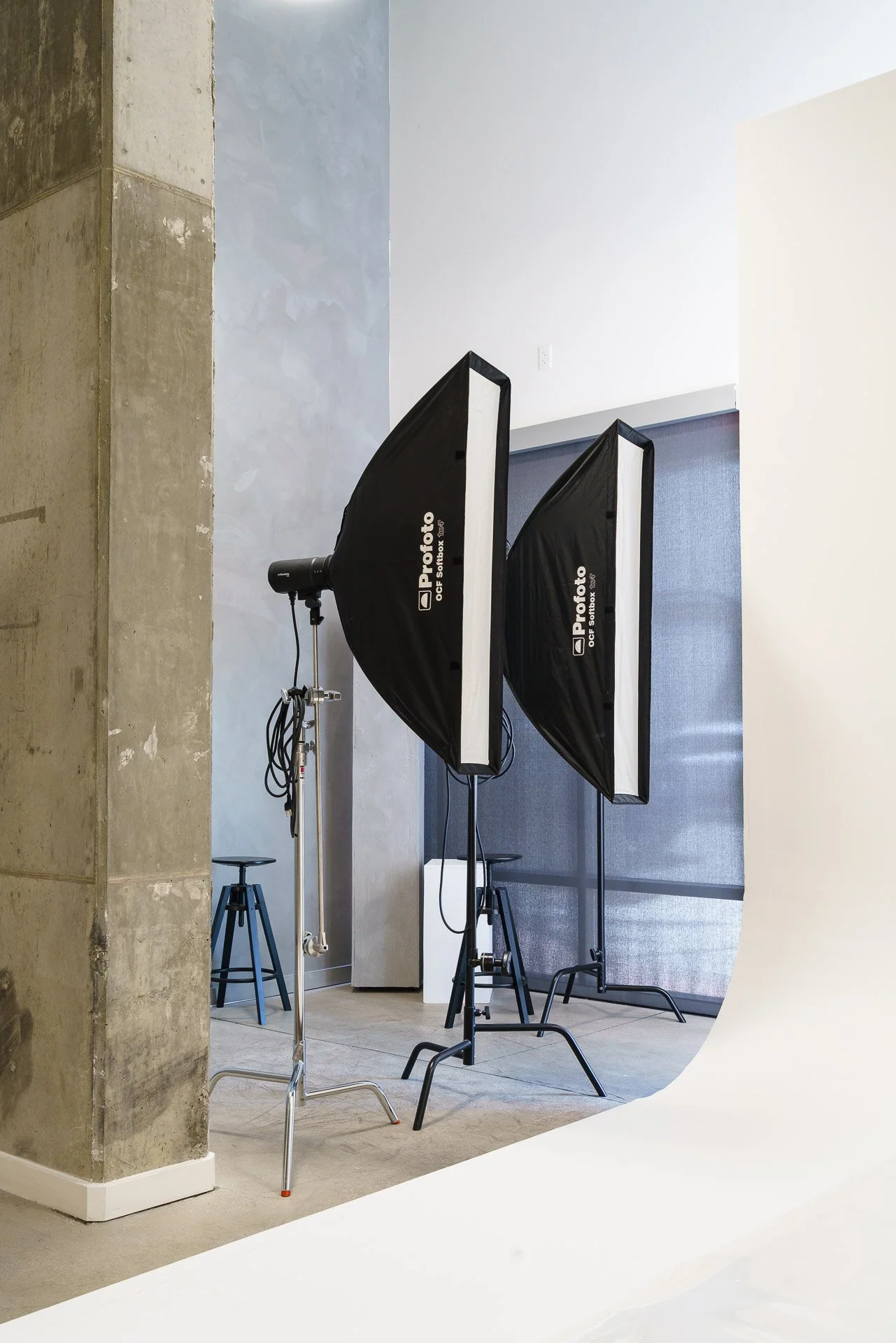 Photography studio with two black softbox lights on stands, a white backdrop, and a concrete pillar on the left side.